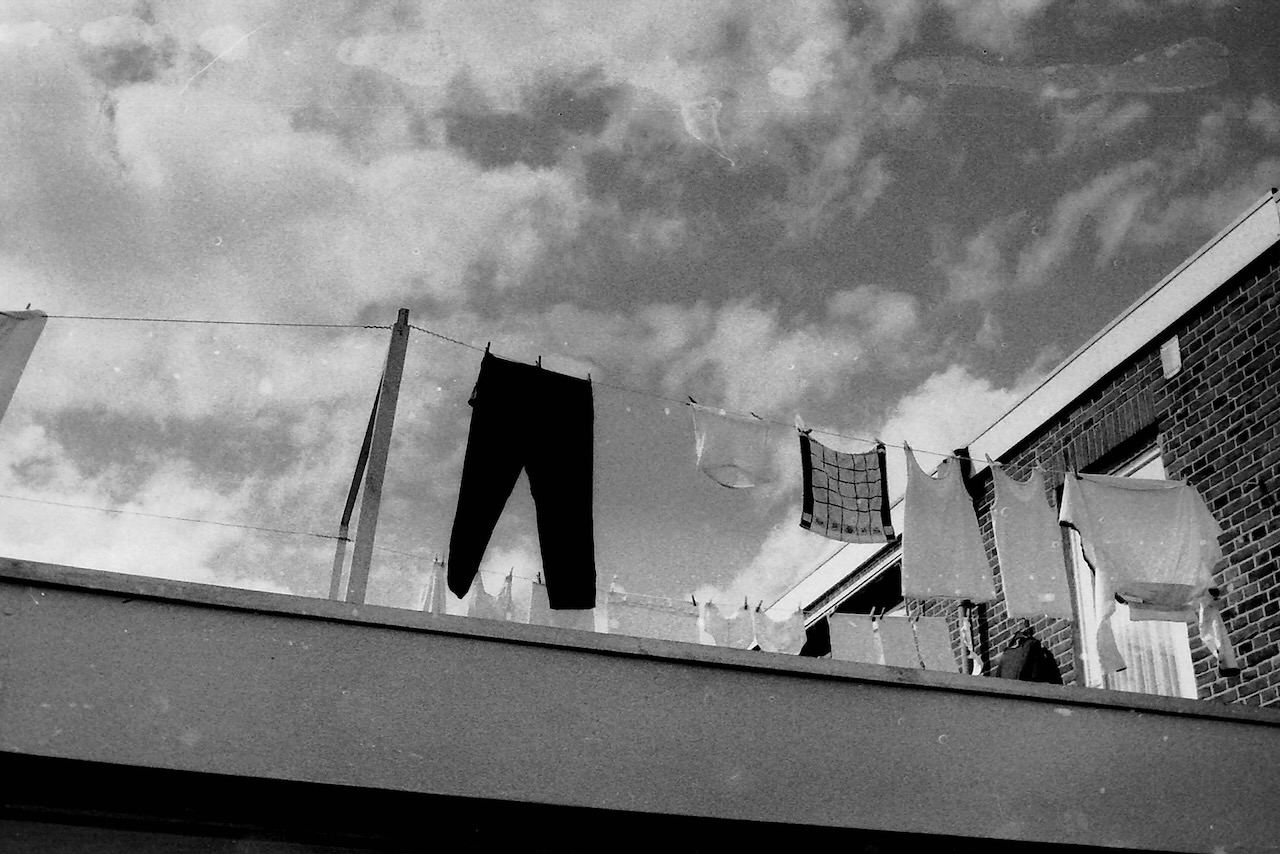 Black and white picture of laundry hanging on a washing line the clouds have summer pattern and the picture is taken from a low angle, the clothes seem to be from a older gentleman. It almost appears that some clothes are floating in the sky.