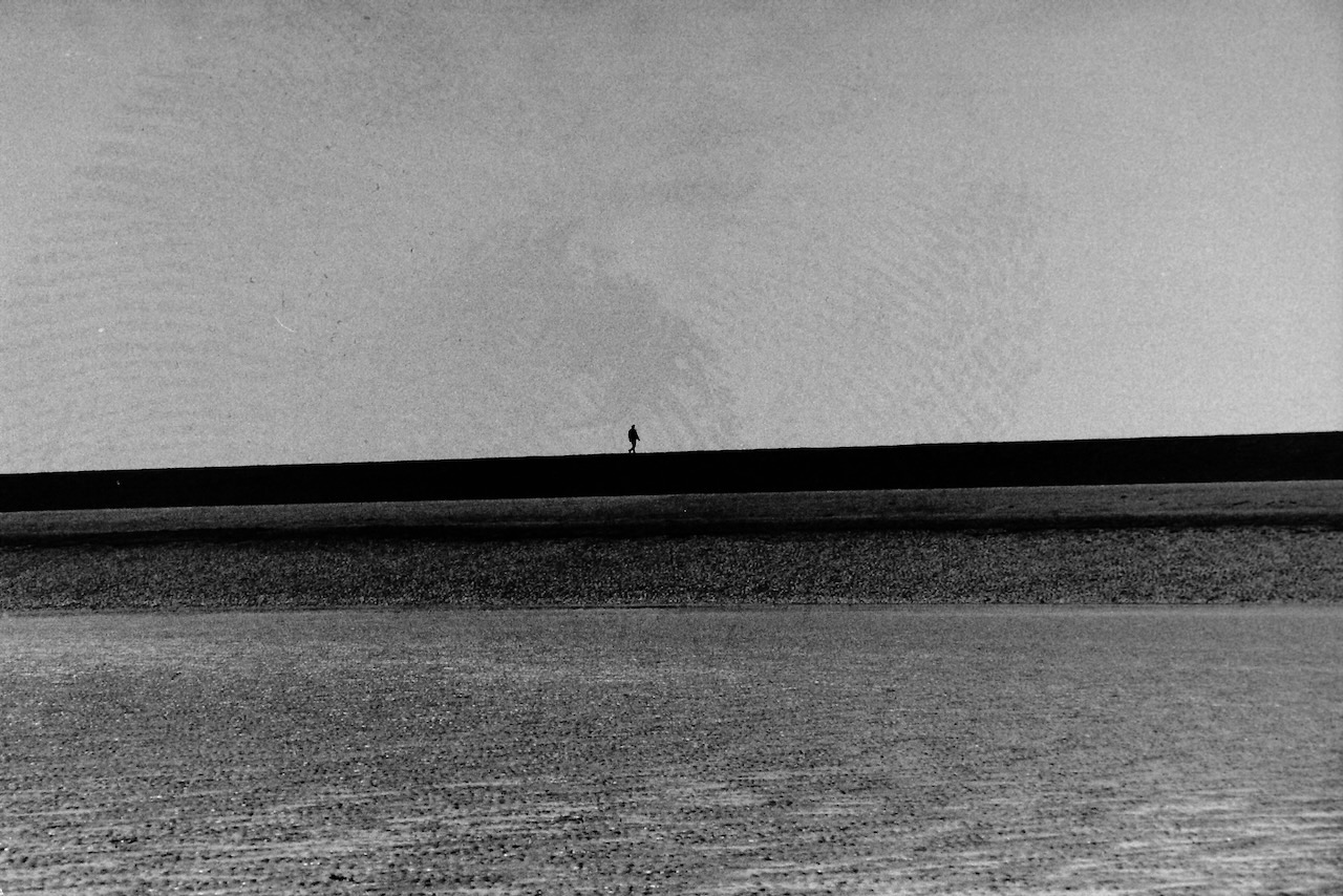 A landscape picture of a dyke, long and flat, but taken askew the horizon seems to be going up. A small silhouette of a person walking is in the middle of the photo. The sky contains a fingerprint, a remainder of the development process. 