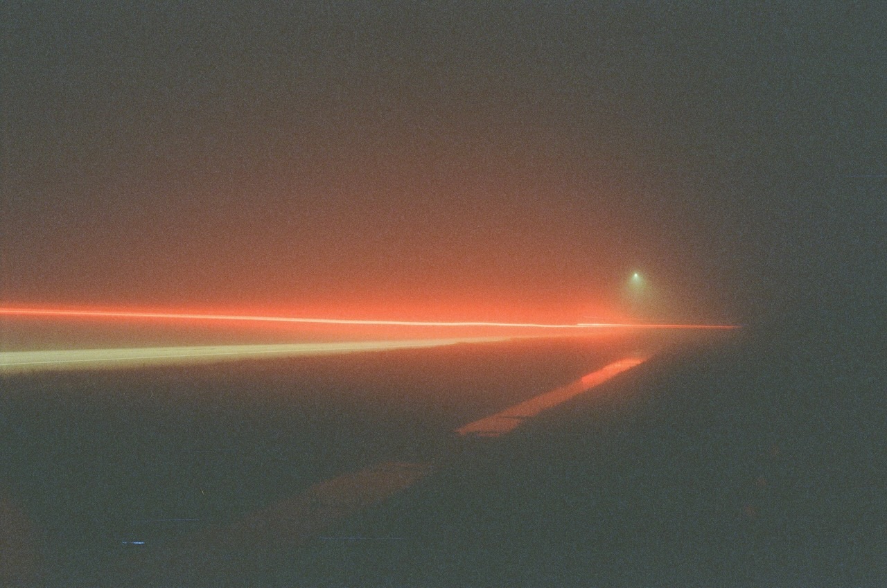 A picture of a red lightbeam traveling along a road. The road is clearly marked with white paint. In the far distance there is a small streetlight. The red laser light seems to brighten the picture with red neon colors.