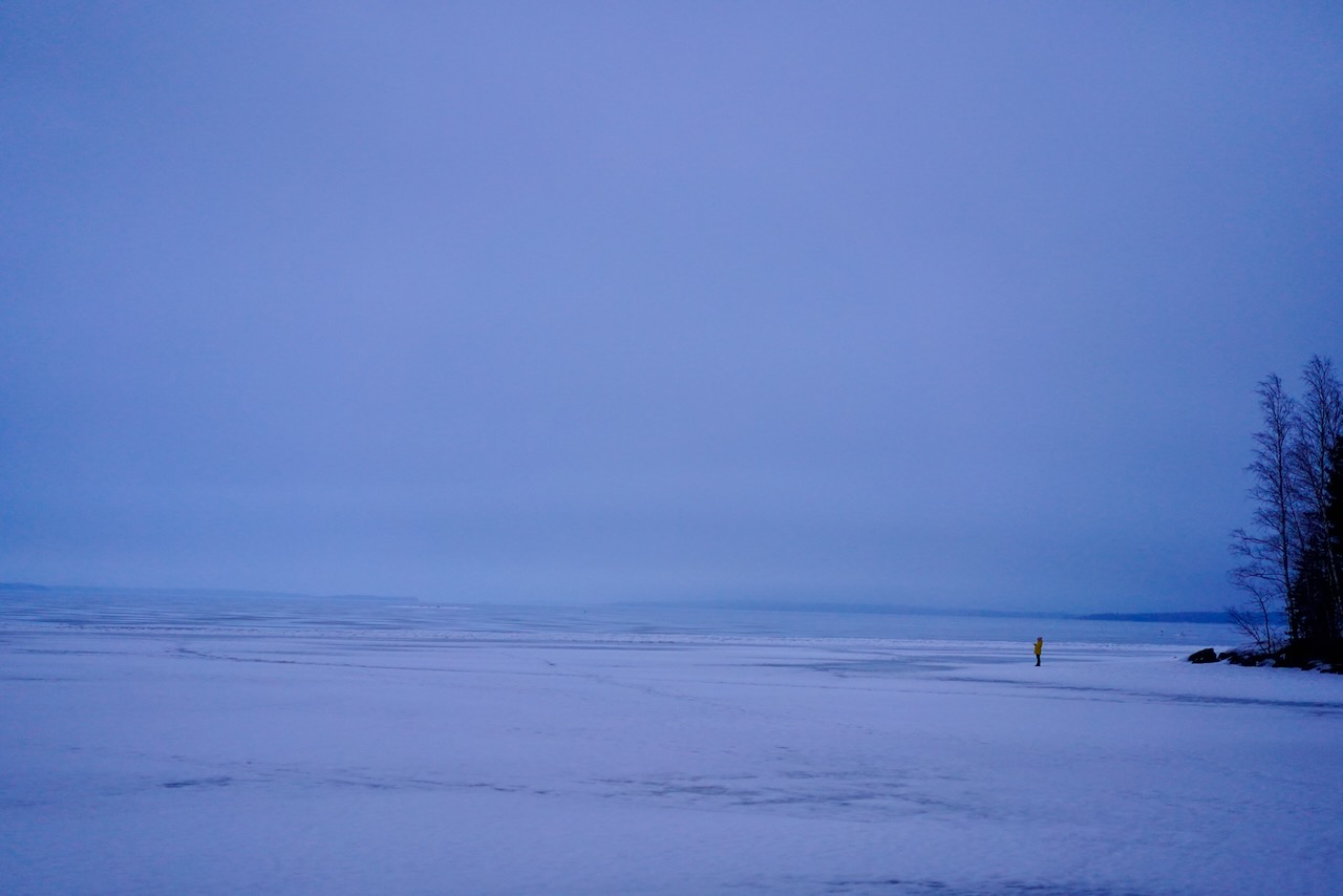 A frozen lake in Finland, washed out blue and white colors. A person wearing a yellow jacket is taking a picture of the lake.