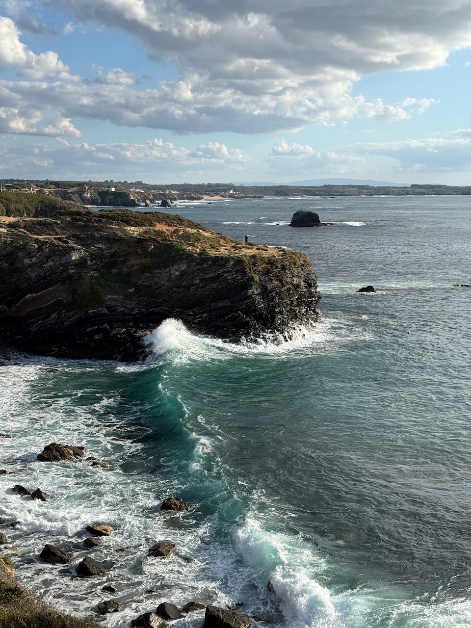 A big rocky cliff in Portugal, one wave is hitting the rock, you can see the outline of the coast. One fisherman is standing on the cliffside, he is far away and seems small. Clouds are stretching far, it seems warm but cold.