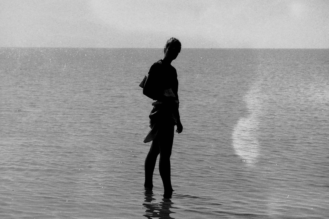 A high contrast picture of a woman standing in the North Sea in low water, the water reaches her ankles. She seems to be one silloette. Her clothes seems to be matching the wavy pattens of the water. She is looking at the person taking the picture. The sea stretches far, and the horizon is flat and almost infinite.