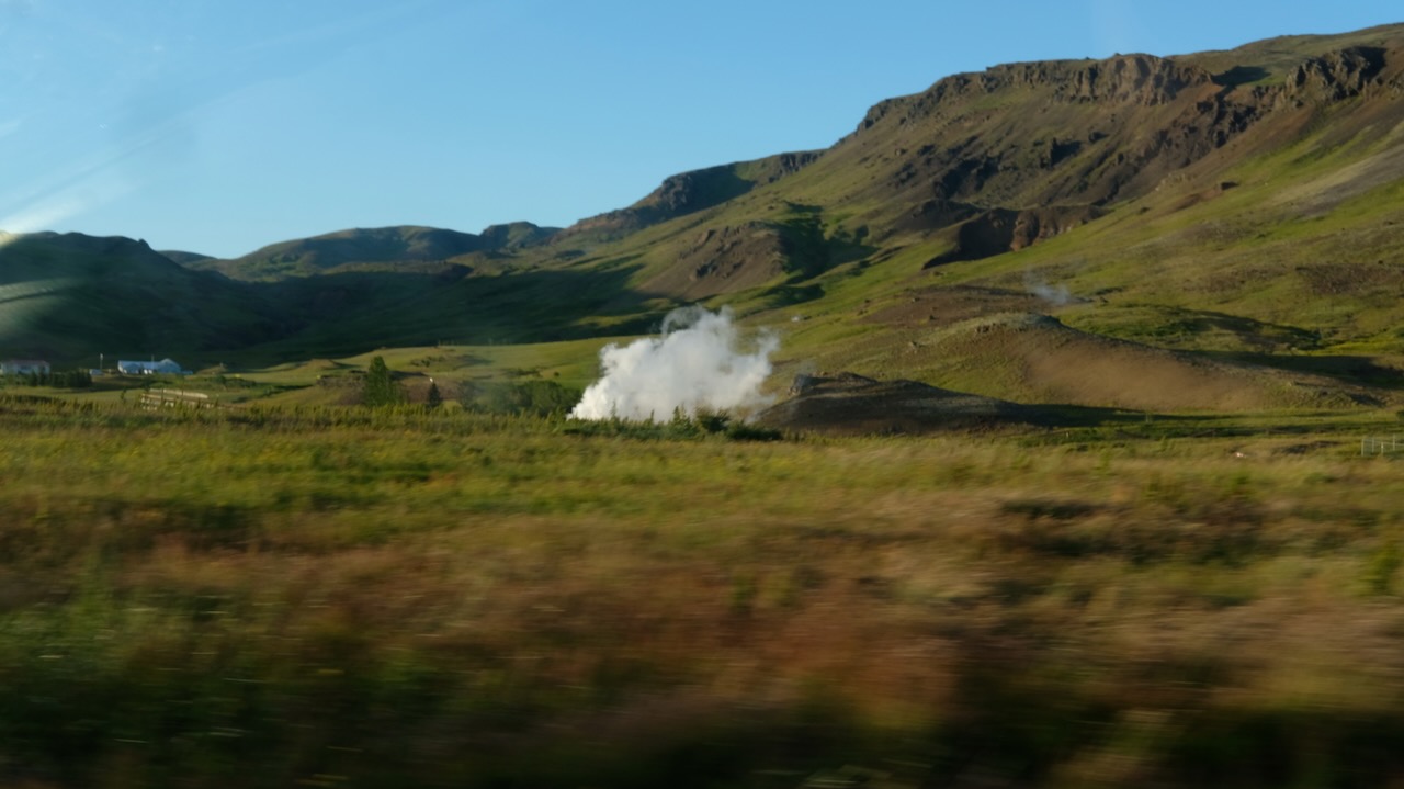 A picture taken from a fast moving car, green mountains are passing by. In the middle there seems to be a steam cloud. The foreground seems to be a high grass of diffrent colours, blurred by the speed of the car.